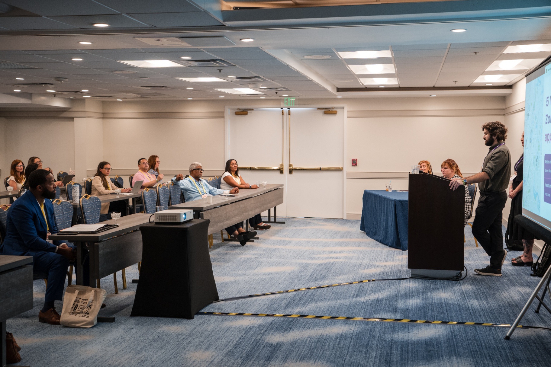 Wide room view of presenters speaking at the front of a Day 1 session