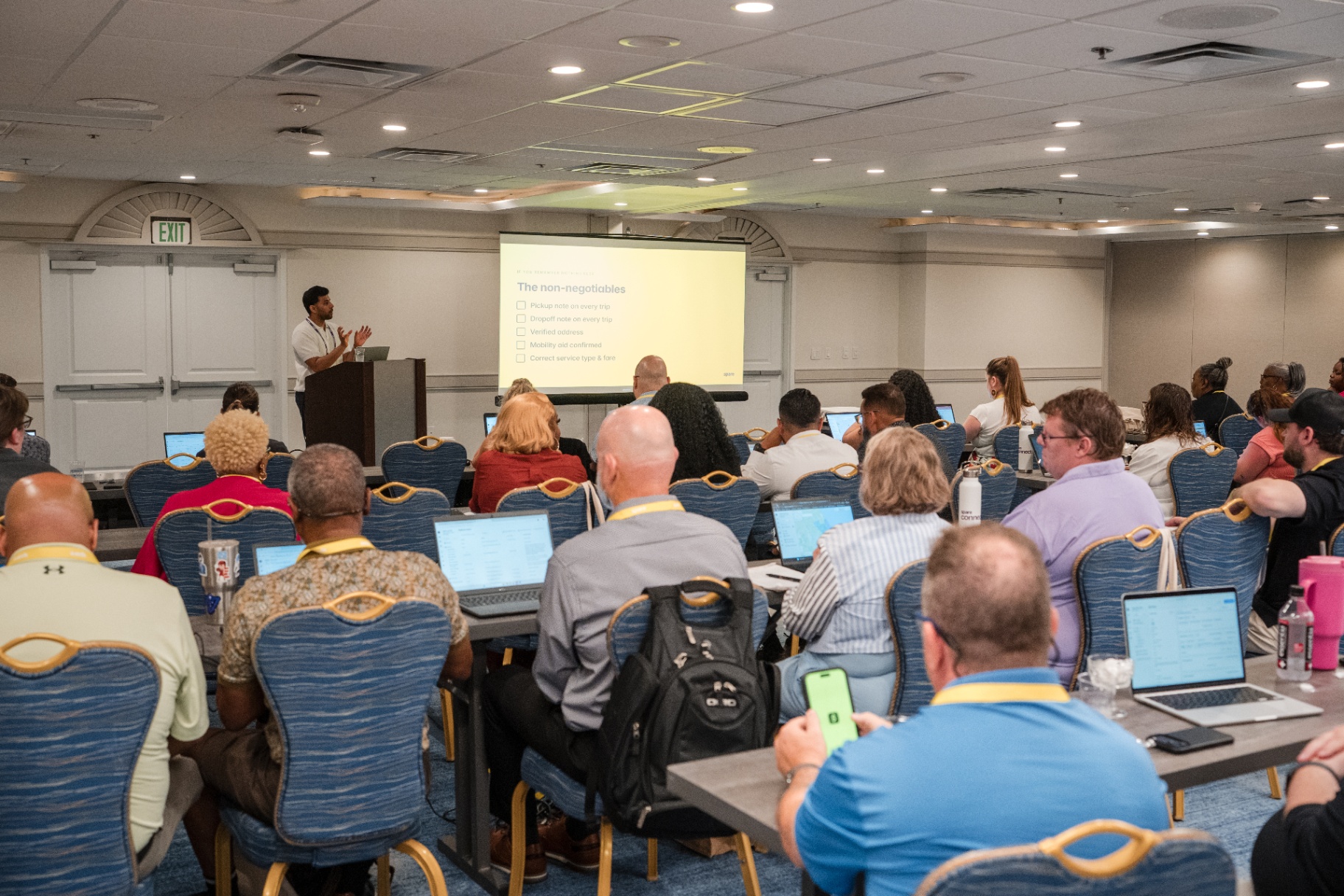 Wide back-of-room view of the speaker presenting the non-negotiables slide